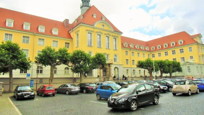 Das gelbe Herforder Rathaus mit rotem Ziegeldach und Turm, davor Platanen und parkende Autos auf dem Rathausplatz